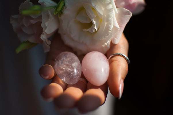 Female hands holding two yoni eggs for vumfit, imbuilding or meditation are made from pink quartz and transparent violet amethyst with white flowers indoors